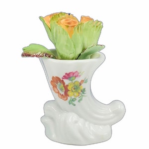 May include: A white porcelain vase shaped like a horn with a floral design. The vase is filled with a single yellow rose with green leaves.