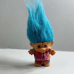 May include: A vintage troll doll with bright blue hair. The doll has a tan face, small blue eyes, and a smiling expression. It is wearing a colorful outfit with red, purple, and yellow accents. The doll is standing upright.