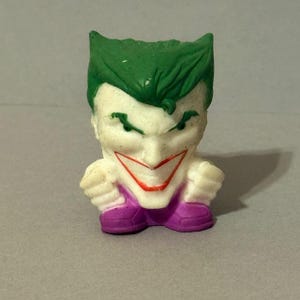May include: A small, stylized figurine of the Joker, a DC Comics supervillain. The figure has a white face with green hair, a red smile, and purple pants and shoes. The figure is in a squatting position with its fists clenched.