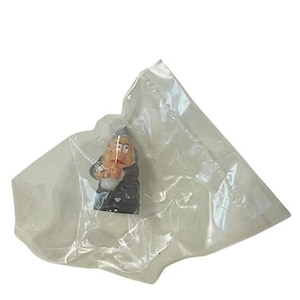 May include: A small ceramic figurine of a gnome wearing a black hat and a black coat. The gnome has a white beard and a brown face.