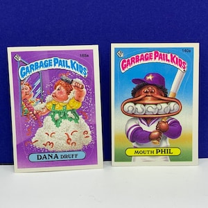 May include: Two Garbage Pail Kids trading cards. One card features "DANA DRUFF" with a character in a yellow dress and white fluff. The other card features "MOUTH PHIL" with a baseball bat and baseballs in his mouth.