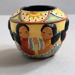 May include: A small, round, yellow ceramic pot with a black interior. The pot features two figures in traditional clothing, with black hair and holding hands. The top rim has a sun and geometric design in orange, blue, and yellow.