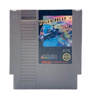 May include: A gray Nintendo Entertainment System game cartridge with a black label. The label features a helicopter flying over a body of water and the text "TIGER-HELI". The label also includes the text "AKlaim entertainment, inc" and "Made in Japan NES-TI-USA".
