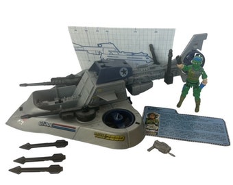 May include: A gray and blue toy helicopter with a star on the side. The helicopter is part of a larger toy vehicle, which is a gray and white hovercraft. The hovercraft has a large cannon on the front and three missiles on the side. The helicopter is attached to the hovercraft by a metal arm. The hovercraft has the words "G.I. JOE" and "WARRIOR FORCE 2000" printed on it. The toy is accompanied by a green toy soldier with a gun and a blue and white instruction sheet.