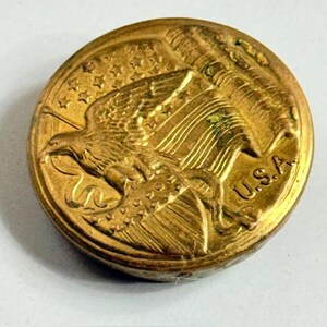 May include: A gold-colored button with an embossed design featuring an eagle, stars, and a flag. The word "U.S.A." is visible. The button has a slightly worn appearance, suggesting it is vintage or antique.