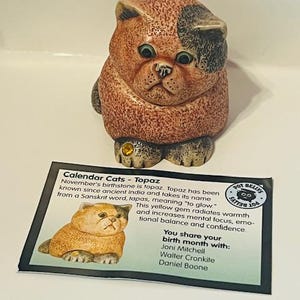 Puede incluir: Una figurita de cerámica de un gato con pelaje naranja y negro. El gato está sentado con la cabeza hacia abajo y las patas metidas debajo de su cuerpo. La figurita forma parte de una colección llamada "Calendar Cats" y está asociada a la piedra de nacimiento topacio. La tarjeta incluye el texto "Calendar Cats - Topaz", "La piedra de nacimiento de noviembre es el topacio. El topacio se conoce desde la India antigua y toma su nombre de la palabra sánscrita tapas, que significa "brillar". Esta gema amarilla irradia calidez y aumenta la concentración mental, el equilibrio emocional y la confianza. Compartes tu mes de nacimiento con: Joni Mitchell, Walter Cronkite, Daniel Boone."