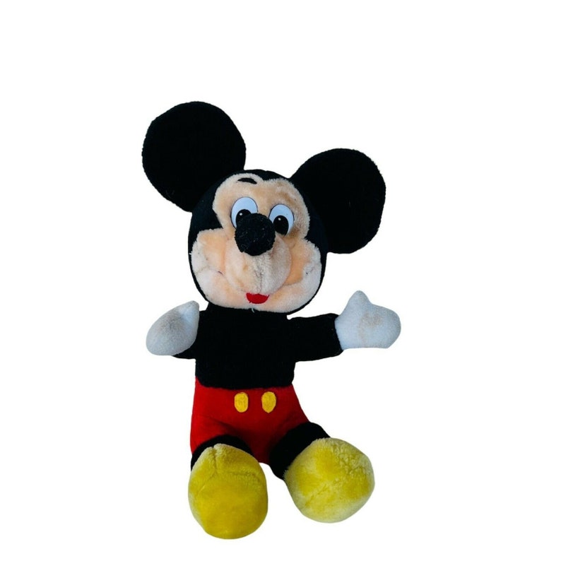 Puede incluir: Un juguete de peluche de Mickey Mouse con orejas negras, un cuerpo negro, pantalones cortos rojos con botones amarillos y zapatos amarillos. El juguete tiene una cara sonriente con ojos negros y una nariz negra.
