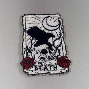 以下が含まれることがあります： 黒いカラス、ドクロ、三日月、赤いバラ2輪が刺繍されたパッチ。下部には「DEATH」の文字。白地で、使い古したようなヴィンテージ感のあるデザイン。