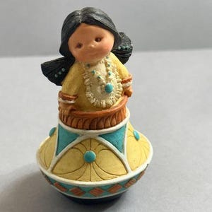 May include: A figurine of a Native American girl with black hair and a yellow dress. The figurine has a turquoise necklace and is standing on a yellow and turquoise base with a decorative pattern. The figurine is a collectible item.