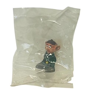 May include: A small, green and brown figurine of a character wearing a black hat and a green jacket. The figurine is sitting down with its legs crossed and its arms folded. The figurine is packaged in a clear plastic bag.