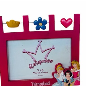 Può includere: Cornice per foto in plastica rosa a tema principessa. Presenta una corona rosa, la parola "Princess" e una cornice per foto 10x15 cm. La cornice è decorata con una corona gialla, un fiore blu e un cuore rosa. La parte inferiore della cornice dice "Disneyland".