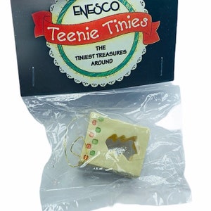 May include: A small, cream-colored ceramic ornament shaped like a house with a cut-out Christmas tree design. The ornament has a gold string for hanging and is packaged in a clear plastic bag with a card that reads "Enesco Teenie Tinies The Tiniest Treasures Around".