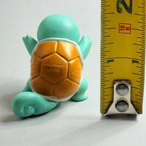 Puede incluir: Figura turquesa de una criatura parecida a una tortuga con caparaz&oacute;n naranja. La figura mide aproximadamente 5 cm de alto. La concha tiene un dise&ntilde;o texturizado. Es un art&iacute;culo de colecci&oacute;n.