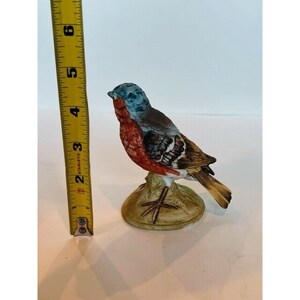 K&ouml;nnte beinhalten: Eine Porzellanfigur eines kleinen Vogels mit blauen, roten, braunen und gelben Markierungen. Der Vogel sitzt auf einem kleinen, braunen Sockel.