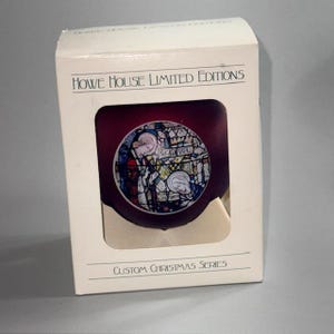 May include: A Christmas ornament in a box. The ornament is a deep red color with a stained-glass design featuring religious figures. The box is white with the text "Howe House Limited Editions" and "Custom Christmas Series".