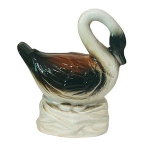 Puede incluir: Una figurita de cisne de cerámica con marcas marrones y negras, posada sobre una base blanca. El cisne tiene un cuello largo y curvo y un pequeño pico.