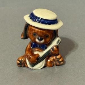 Puede incluir: Figura de cerámica de un perro marrón con un sombrero blanco con una banda azul y una pajarita azul. El perro sostiene un banjo. La figura tiene un acabado brillante y está sobre un fondo gris.