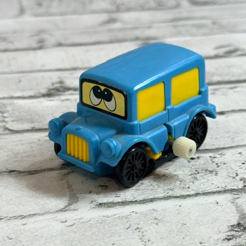 May include: A small blue plastic toy car with a wind-up mechanism on the side. The vehicle features a yellow front grille, yellow side windows, and a windscreen displaying two large cartoon eyes with black pupils looking towards the upper left.