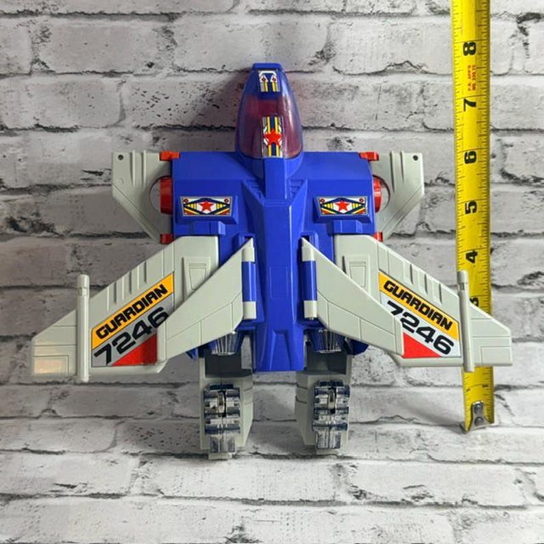 May include: A blue and light gray plastic toy jet with red accents and a translucent cockpit canopy. The wings feature yellow decals with the text GUARDIAN 7246. A yellow measuring tape is positioned vertically, showing a height of 8 inches.