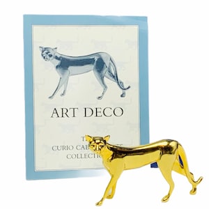 May include: A gold-colored figurine of a stylized cat, standing on a white surface. The figurine is part of an Art Deco collection. The text "Art Deco" is printed on a blue card behind the figurine.