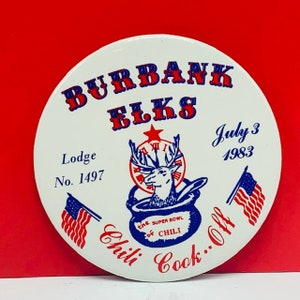 May include: A white button with a red, white, and blue design. It says "Burbank Elks Lodge No. 1497" and "The Super Bowl of Chili" with a picture of an elk in a pot. It also says "July 3, 1983" and "Chili Cook-Off".
