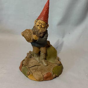 May include: A whimsical garden gnome figurine with a red pointed hat, holding a large nut. The gnome has a long, brown beard and is dressed in a blue jacket and brown pants. The base is decorated with leaves and acorns.