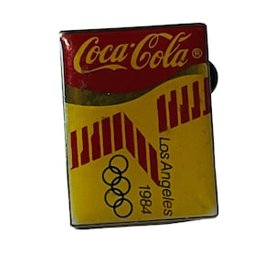 May include: A red and yellow Coca-Cola pin with the Olympic rings and the text "Los Angeles 1984".