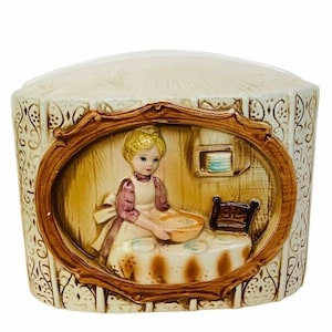 May include: A ceramic napkin holder with a raised oval scene of a woman in a kitchen setting. The woman is wearing a pink and white dress and is holding a bowl of flour. The scene is framed in brown and the holder has a white background with a brown floral pattern.