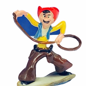 May include: A small, colourful figurine of a cowboy with a lasso. He is wearing a red hat, yellow shirt, blue waistcoat, and brown trousers. The figurine is made of painted metal and is about 3 inches tall.