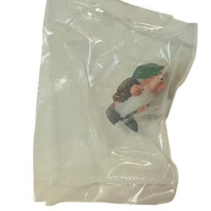 May include: A small plastic figurine of a person wearing a green hat and holding a white object. The figurine is packaged in a clear plastic bag.