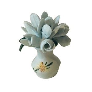 May include: A small white ceramic vase with a floral design painted on the side. A light blue ceramic flower arrangement is in the vase.