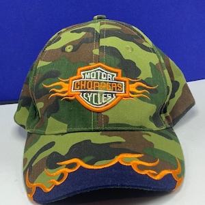 May include: A green, brown, and black camouflage baseball cap with an orange and white embroidered patch that says "Motor Choppers Cycles". The cap has an orange flame design on the brim.