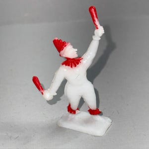 May include: A small, white plastic clown figurine with red detailing. The clown is holding two red batons, wearing a red hat, collar, and shoes. The figurine stands on a white base, against a neutral background.