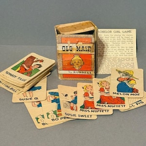 May include: An antique "Old Maid" card game by Russell. The box is orange and white with the game title and a cartoon illustration. Several cards are visible, featuring cartoon characters with names like "Tomboy Tillie" and "Melon Moe."