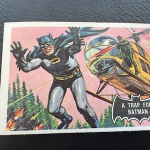 Könnte beinhalten: Eine Vintage-Comic-Sammelkarte mit Batman in einem schwarzen Anzug, der durch die Luft fliegt, mit einem gelben Hubschrauber im Hintergrund. Die Karte trägt den Text "A Trap For Batman" in einem schwarzen, fledermausförmigen Banner.