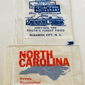 May include: Two white paper sugar sachets. The top sachet has blue text that reads "Maxwell House Coffee Vicki Villa Restaurant Air Conditioned Serving the South's Finest Food Elizabeth City, N.C.". The bottom sachet has red text that reads "North Carolina" and a blue outline of the state of North Carolina. Below the state outline is the text "Variety Vacationland".