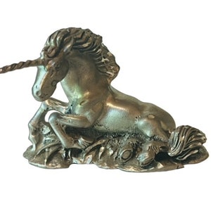 May include: A silver pewter figurine of a unicorn lying down with its head turned to the side. The unicorn has a long, spiraled horn and a flowing mane and tail.