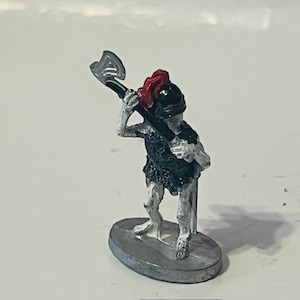May include: A white painted miniature skeleton figure with a black chainmail shirt and a red and black helmet holding a silver axe. The figure is standing on a gray base.