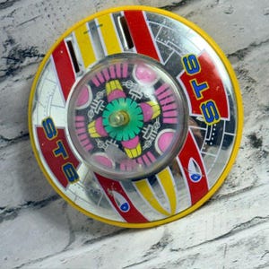 May include: A round, vintage-style tin toy spinning top featuring a yellow outer rim, red and white striped accents, and the text ST6 printed in blue. The centre has a clear plastic dome covering a colourful, multi-patterned rotating disc.