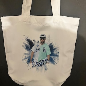 May include: White canvas tote bag with a blue and white graphic print of a person wearing a baseball cap, sunglasses, and a baseball jersey with the text "Los A" and "LA" on it. The text "Dodgers" is printed in blue below the person.