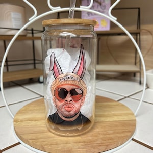 May include: A clear glass tumbler with a bamboo lid and a straw. The tumbler has a cartoon image of a person wearing a hat, sunglasses, and bunny ears. The text "#LOMDLG" is on the hat.