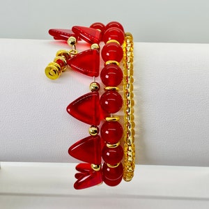 Puede incluir: Tres pulseras apiladas. Una pulsera presenta cuentas rojas triangulares y redondas con detalles dorados. La segunda pulsera tiene cuentas redondas rojas con detalles dorados. La tercera pulsera está hecha de pequeñas cuentas doradas. Las pulseras están sobre una superficie blanca.