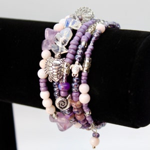 Puede incluir: Una pulsera de múltiples hilos con una mezcla de cuentas moradas, rosas y plateadas. La pulsera presenta varios dijes, incluyendo colgantes de tortugas y conchas marinas. Las cuentas son de diferentes tamaños y formas.