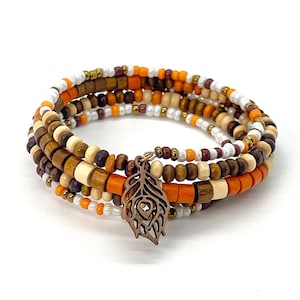 Könnte beinhalten: Ein Wickelarmband mit Holzperlen in Braun-, Orange- und Weißtönen, akzentuiert mit kleinen goldenen und weißen Perlen. Ein kupferfarbener Federanhänger hängt am Armband. Das Armband ist so konzipiert, dass es sich mehrfach um das Handgelenk wickelt.