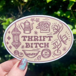 May include: Oval sticker with a tan background and maroon illustrations of a teapot, telephone, and other vintage items. The words "THRIFT BITCH" are in the center. The sticker is held up against a background of green leaves.