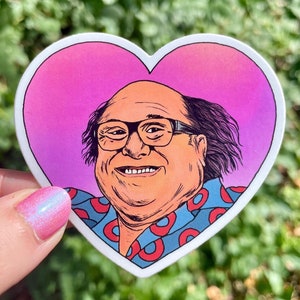May include: A heart-shaped sticker with a cartoon illustration of a man with glasses wearing a blue shirt with red donut patterns. The background is a pink to purple gradient.