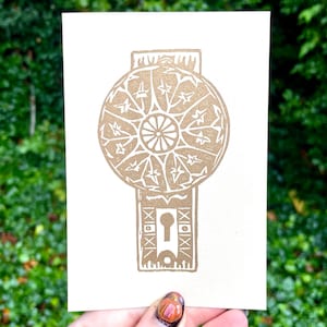 May include: A cream-colored card featuring a gold-colored illustration of a vintage doorknob design. The design includes a circular element with star and floral patterns, and a keyhole. The card is held against a backdrop of green foliage.