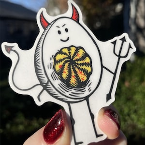 May include: A white egg with a devil costume, red horns, and a tail. The egg has a smiling face and is holding a pitchfork. The egg's yolk is visible and has a red and yellow swirl pattern.