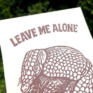 以下が含まれることがあります： 茶色のアルマジロのイラストと「LEAVE ME ALONE」の文字が描かれたプリント。アルマジロは詳細に描かれています。背景はオフホワイトです。ぼやけた緑色の背景にプリントされています。