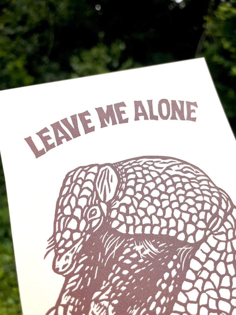 以下が含まれることがあります： 茶色のアルマジロのイラストと「LEAVE ME ALONE」の文字が描かれたプリント。アルマジロは詳細に描かれています。背景はオフホワイトです。ぼやけた緑色の背景にプリントされています。